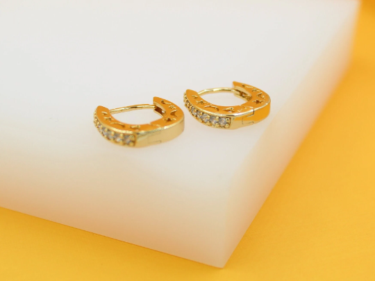 A pair of gold huggie hoop earrings featuring sparkling pave-set stones and celestial cut-outs rests on a white display surface against a vibrant yellow background.