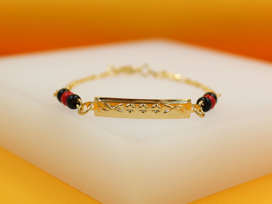 A delicate gold identification bracelet features an engraved rectangular plate flanked by alternating black and red beads resting on a white display surface.