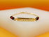 A delicate gold identification bracelet features an engraved rectangular plate flanked by alternating black and red beads resting on a white display surface.
