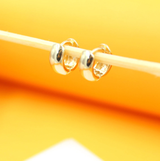 Two shiny gold huggie hoop earrings hang delicately from a wooden stick against a bright yellow background.