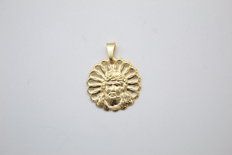 A round gold pendant features an embossed face of Jesus surrounded by a halo-like cut-out border against a plain background.