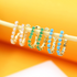 Three pairs of gold hoop earrings adorned with pink, green, and turquoise stones hang in a row on a wooden stick against a warm yellow and orange background.