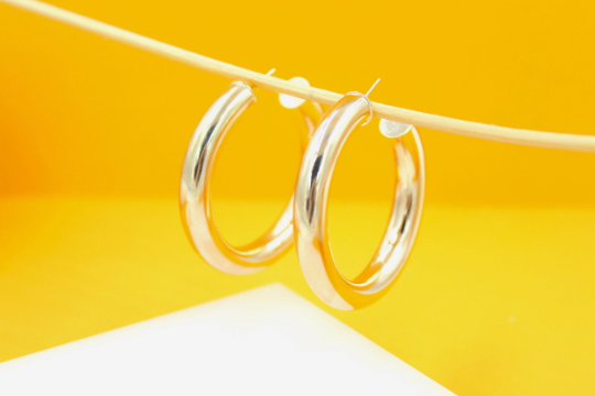 A pair of thick silver hoop earrings hangs from a wooden stick against a bright yellow background.