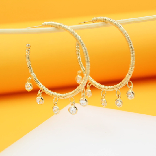 Two gold-plated hoop earrings with textured wire wraps and dangling cubic zirconia charms hang displayed against a bright yellow background.