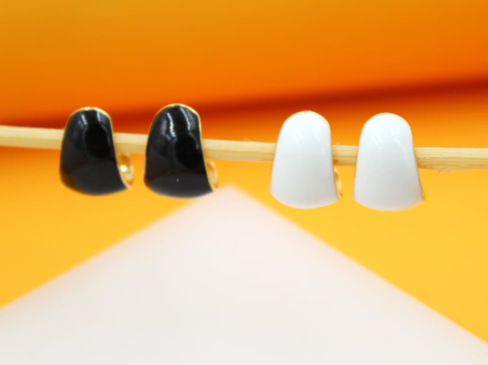 Two pairs of glossy black and white enamel hoop earrings rest on a wooden stick against a bright yellow background.