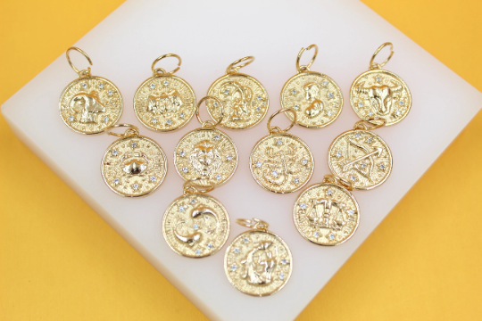 A collection of gold zodiac sign pendants with sparkling crystal accents rests on a white display block against a yellow background.