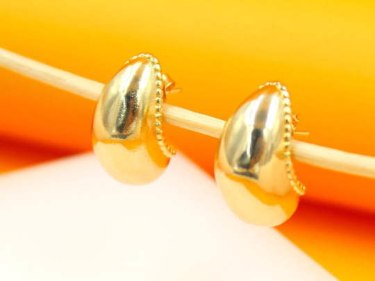 A pair of polished gold teardrop huggie earrings with beaded trim rests on a thin wooden stick against a bright yellow and white background.