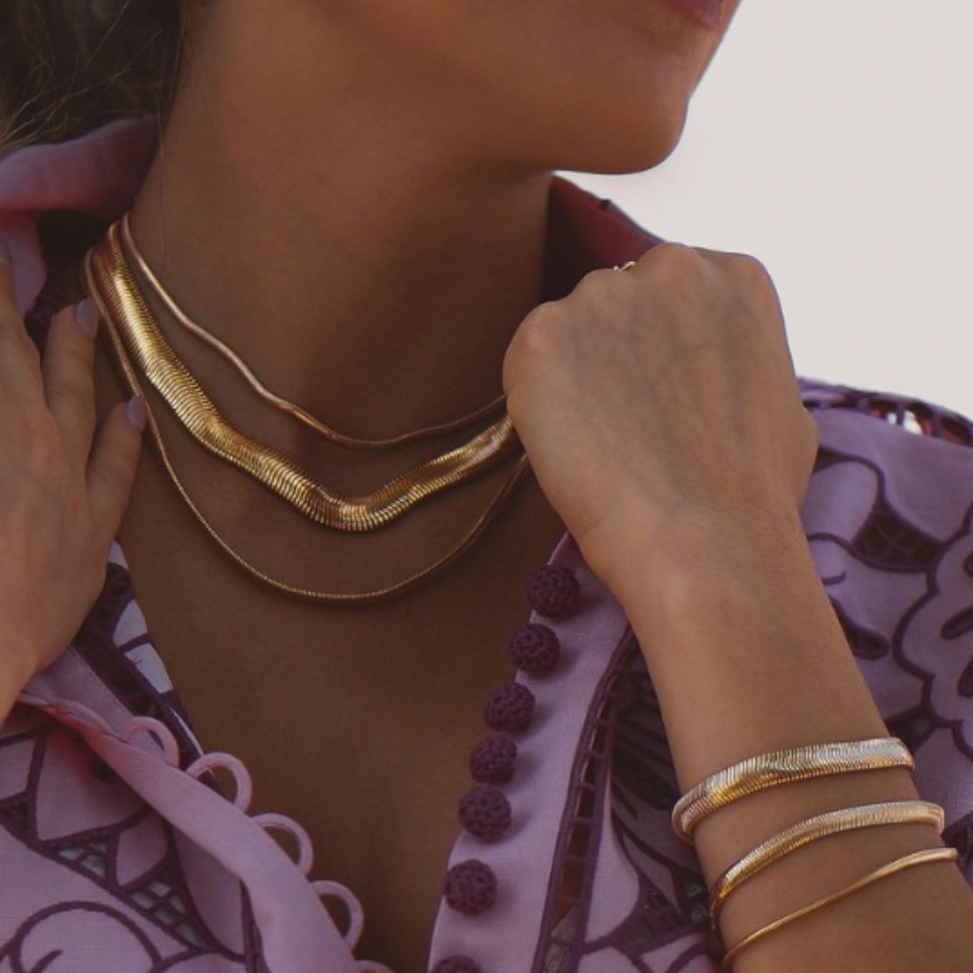 A stylish woman wearing layered gold snake chain necklaces and matching bracelets with a purple embroidered blouse.