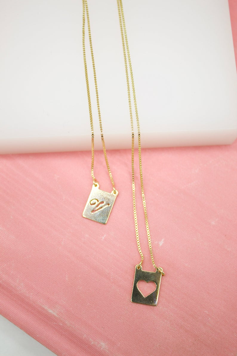Two delicate gold chain necklaces with rectangular pendant charms, one featuring an engraved script letter and the other a cutout heart, rest against a pink surface.