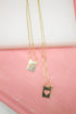 Two gold chain necklaces featuring rectangular pendants with a letter "V" and a heart cutout rest against a pink and white background.