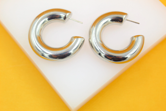 A pair of thick silver hoop earrings rests neatly on a white display stand against a bright yellow background.
