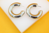 A pair of thick silver hoop earrings rests neatly on a white display stand against a bright yellow background.