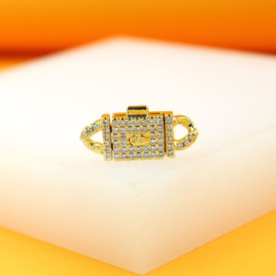A gold jewelry clasp encrusted with sparkling cubic zirconia stones rests on a white display surface.