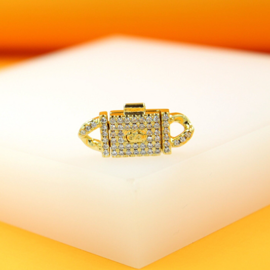 A gold jewelry clasp encrusted with sparkling cubic zirconia stones rests on a white display surface.