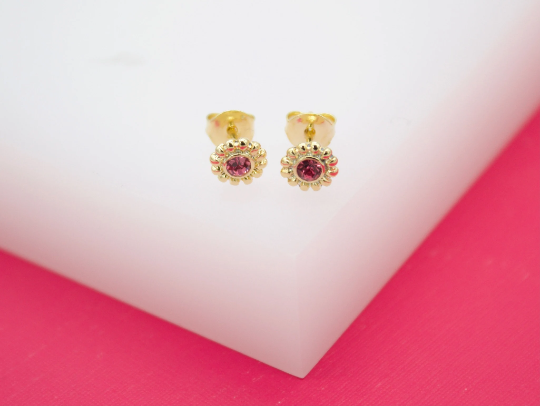 A pair of gold flower-shaped stud earrings featuring sparkling pink crystals rests on a white block against a pink background.