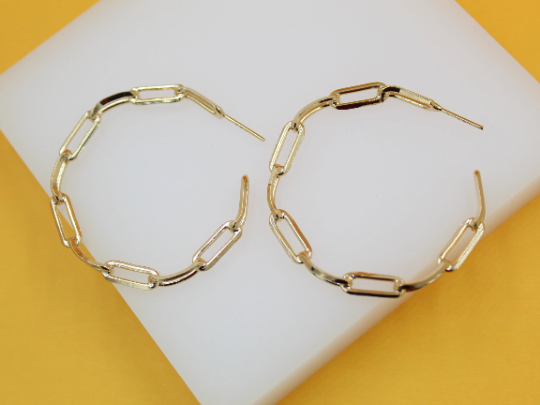 A pair of gold paperclip chain hoop earrings rests on a white display surface against a vibrant yellow background.