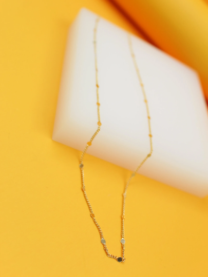 A delicate gold chain necklace with evenly spaced flat circular discs rests diagonally on a white rectangular display against a vibrant yellow background.
