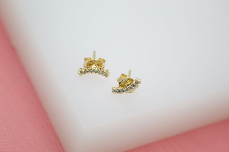 A pair of gold curved stud earrings adorned with sparkling cubic zirconia stones rests on a white surface against a soft pink background.