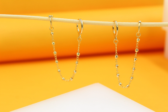 Two delicate gold double-piercing chain earrings hang from a wooden stick against a bright orange and white background.