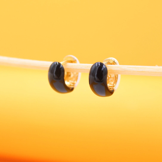 A pair of glossy black and gold hoop earrings hangs delicately on a wooden stick against a warm yellow background.