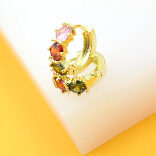 A pair of gold huggie hoop earrings features sparkling multicolor cubic zirconia stones in pink, red, and green against a white and yellow background.