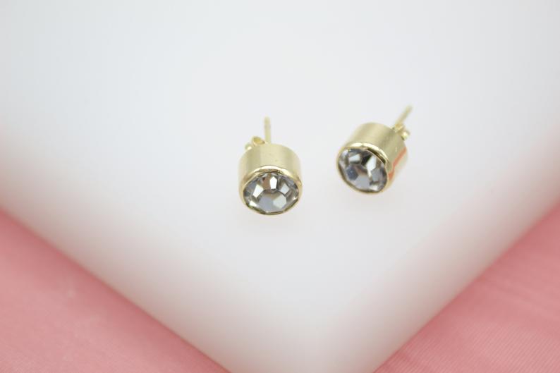 A pair of gold stud earrings featuring sparkling round bezel-set crystals rests on a white display stand.