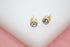 A pair of gold stud earrings featuring sparkling round bezel-set crystals rests on a white display stand.