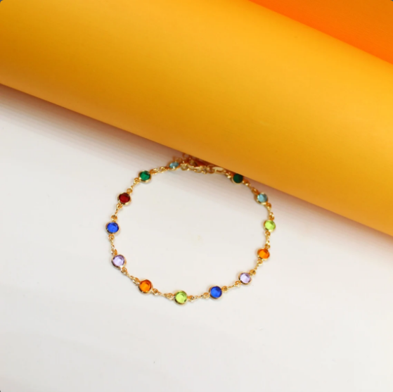 A delicate gold chain bracelet features vibrant multicolored bezel-set crystals resting against a clean white and yellow background.