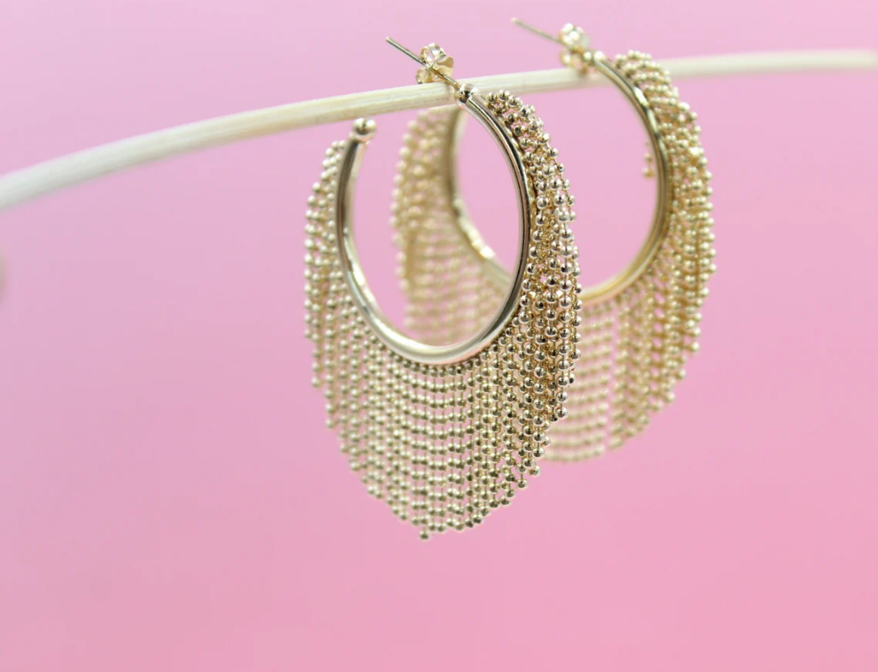 A pair of elegant gold beaded hoop earrings hang delicately from a white twig against a soft pink background.