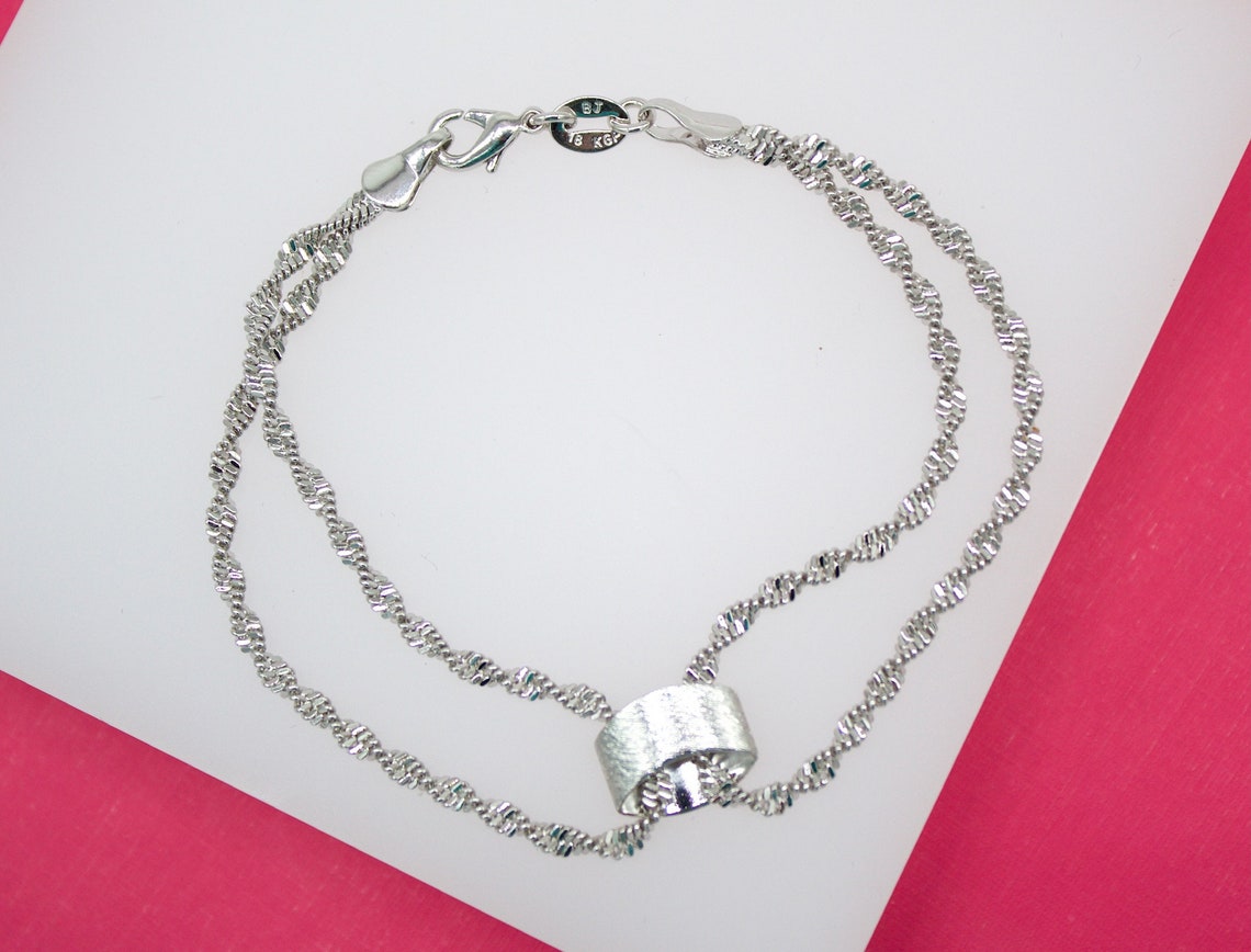 A delicate double-strand silver chain bracelet features a textured cylindrical pendant resting against a white and pink background.