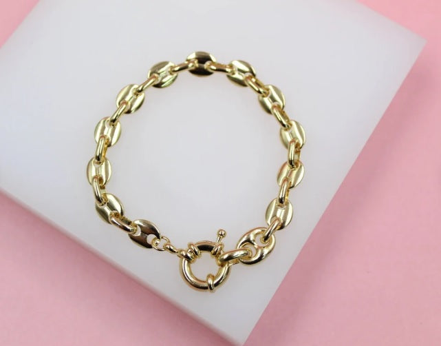 A polished gold coffee bean link bracelet rests on a white display stand against a pink background.