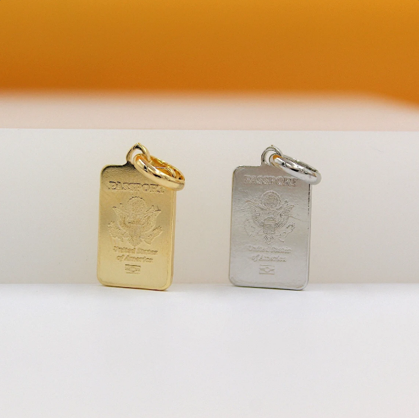 Two shiny rectangular US passport charms, one in gold and one in silver, stand upright side-by-side against a neutral background.