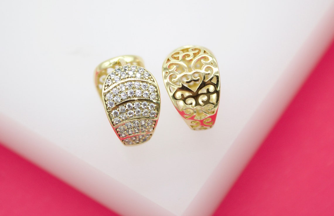 A pair of gold huggie earrings featuring one pavé-set cz diamond half and one filigree cut-out half rests on a white display surface.