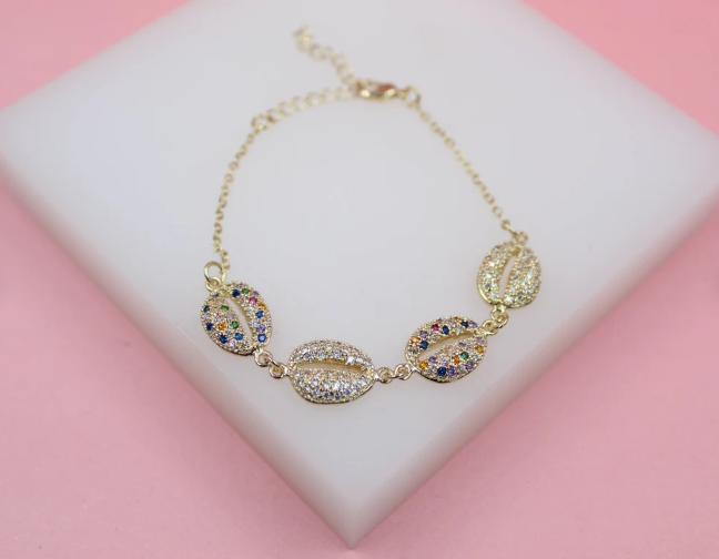 A gold seashell bracelet adorned with sparkling multi-colored cubic zirconia stones rests on a white display stand.