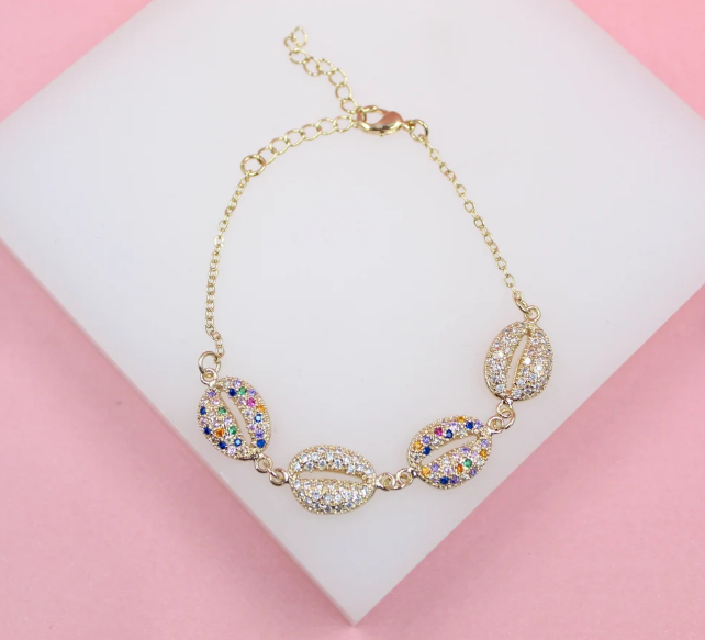 A delicate gold chain bracelet features four glittering shell charms accented with colorful zirconia stones, resting against a white and pink background.