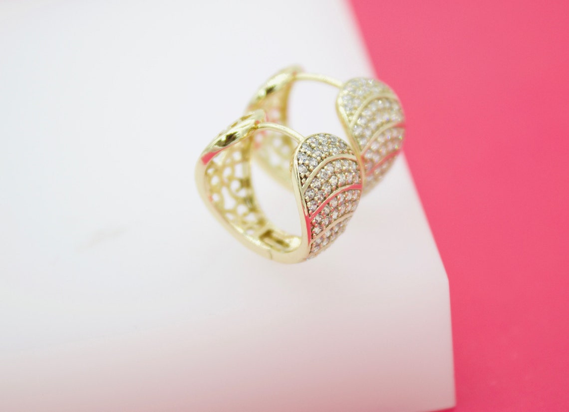 A pair of gold huggie hoop earrings encrusted with sparkling cubic zirconia stones rests on a white display surface against a pink background.