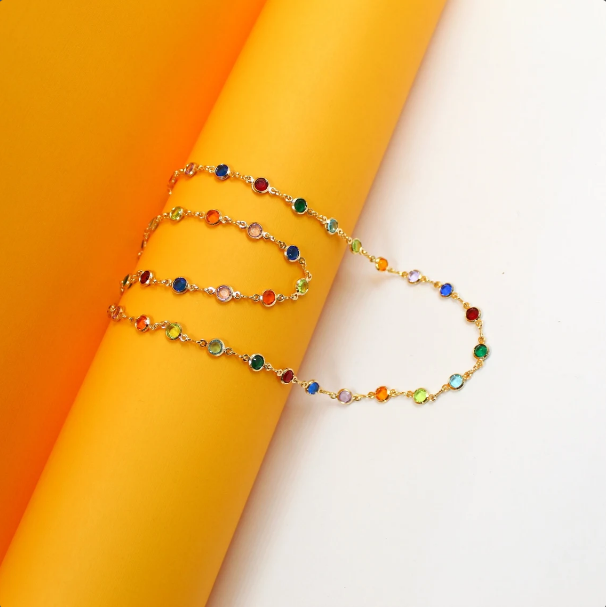 A delicate gold necklace featuring bezel-set multicolored gemstones rests gracefully against a vibrant orange and white background.