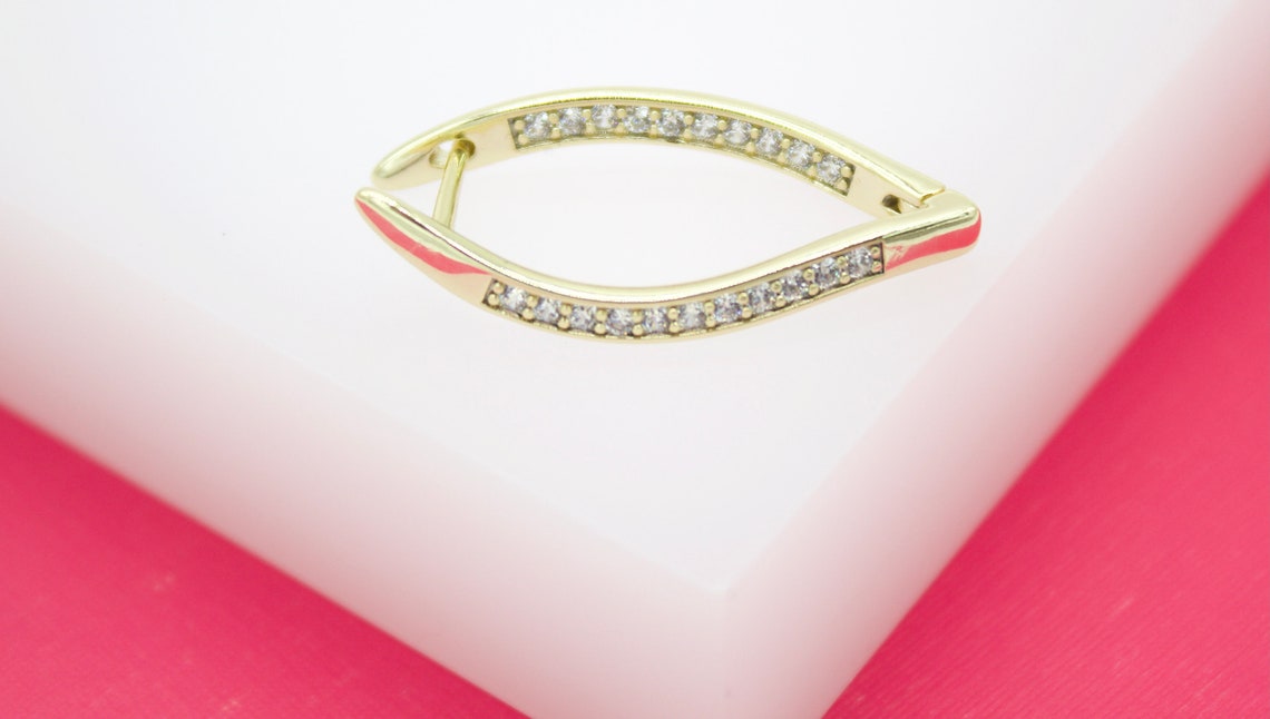 A gold hoop earring featuring an eye-shaped design set with small sparkling cubic zirconia stones rests on a white display surface.