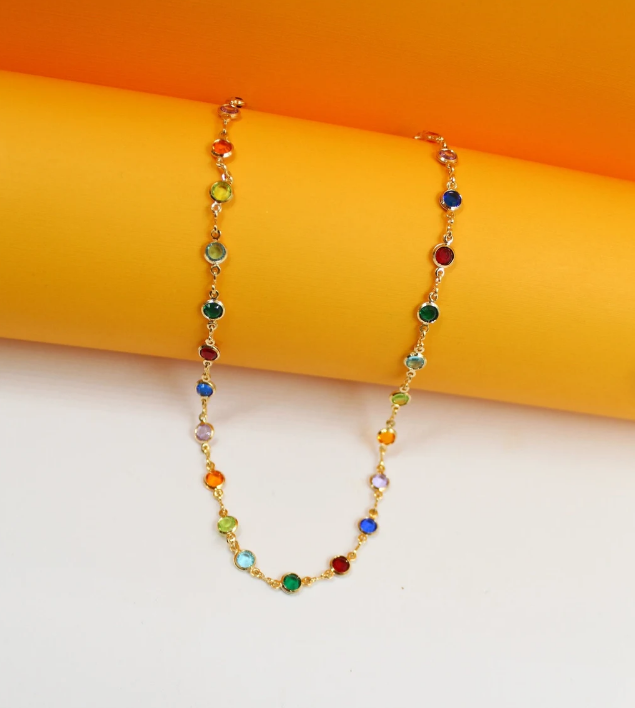 A gold chain necklace drapes gracefully against a vibrant yellow and orange background, showcasing colorful, bezel-set cubic zirconia stones in a rainbow spectrum.