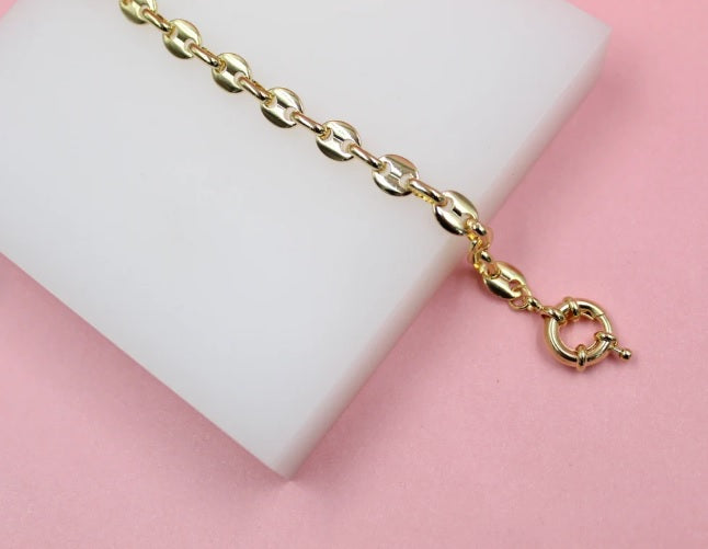 A delicate gold coffee bean chain bracelet rests diagonally on a white display block against a soft pink background.