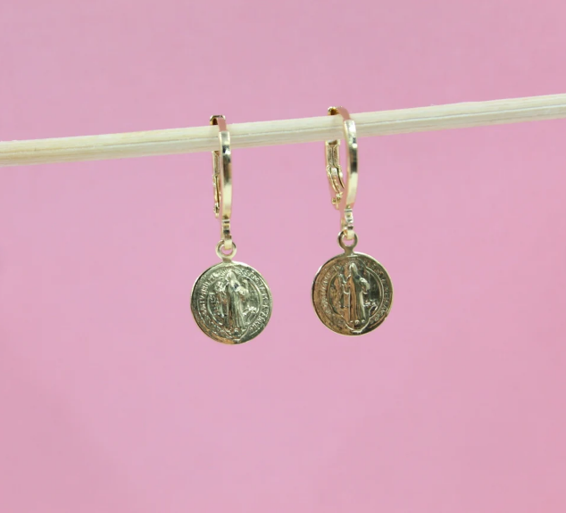 Two gold Saint Benedict medal charm hoop earrings hang neatly from a wooden stick against a soft pink background.
