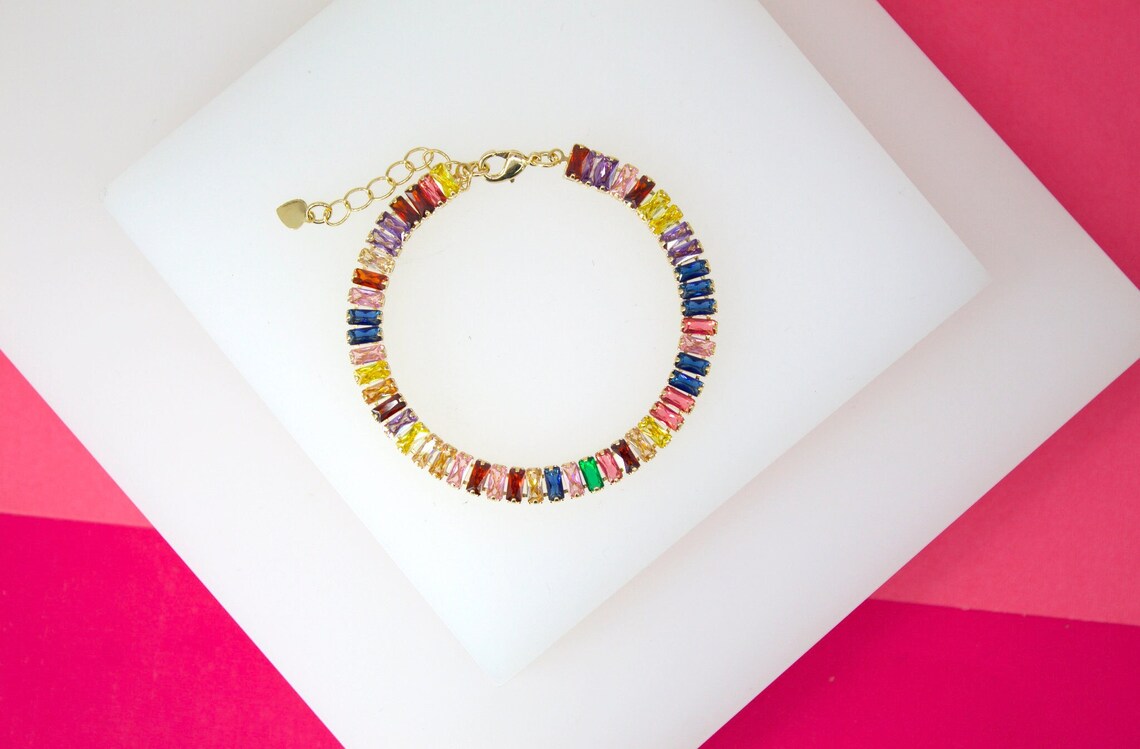 A vibrant gold tennis bracelet features multicolored baguette-cut crystals displayed against a white and pink background.