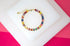 A vibrant gold tennis bracelet features multicolored baguette-cut crystals displayed against a white and pink background.