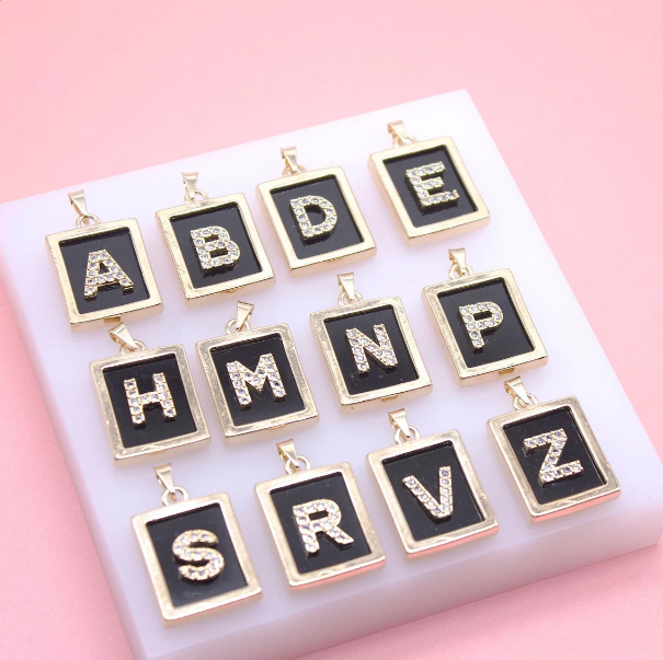 A collection of gold rectangular alphabet pendants featuring sparkly rhinestone initial letters on a black background is arranged neatly on a white display block against a soft pink backdrop.