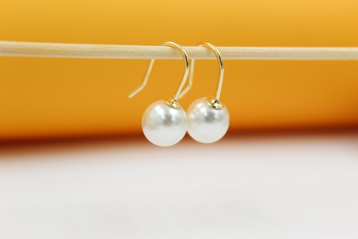Two elegant gold-plated hook earrings featuring lustrous white pearl drops hang gracefully from a wooden stick against a warm yellow and white backdrop.