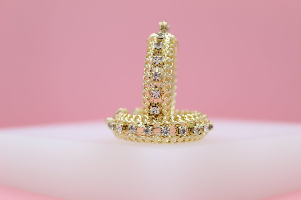 A pair of intricate gold hoop earrings embellished with small sparkling diamonds rest on a white surface against a soft pink background.