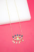 A gold-plated evil eye pendant necklace features a central yellow crystal surrounded by clear rhinestones, blue marquise stones, and dangling gold teardrops against a bright pink background.