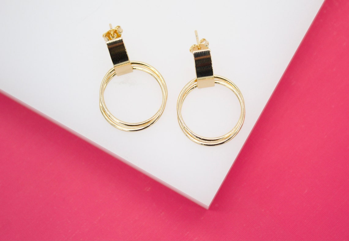 A pair of elegant gold geometric earrings rests on a white display against a vibrant pink background.