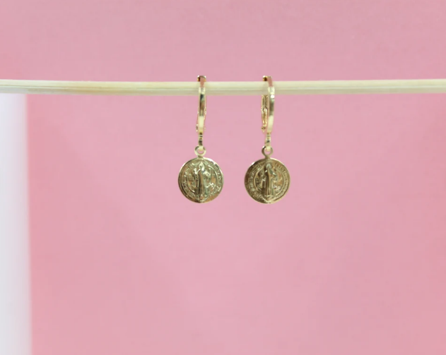A pair of gold coin hoop earrings hangs delicately from a wooden stick against a soft pink background.