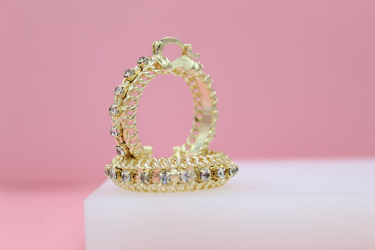 A pair of ornate gold hoop earrings encrusted with sparkling rhinestones rests on a white display block against a soft pink background.